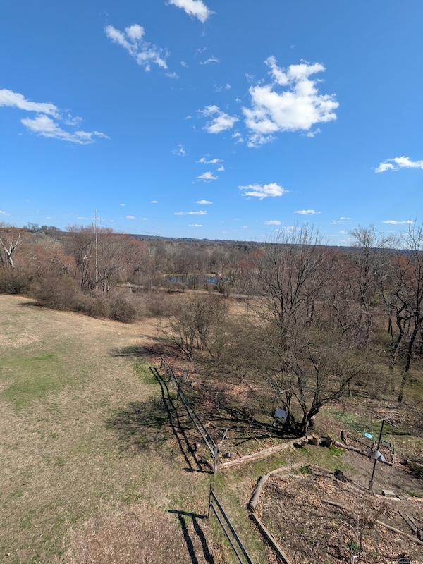 Photo taken from a height, overlooking muddy spring fields, bare trees, and bird feeders. The sky is blue, with a smattering of clouds, and that blue is reflected in water visible through the trees, just beyond the clear path surrounding railroad tracks.